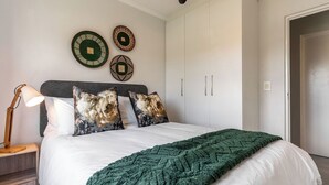 A bedroom with a bed, a nightstand, a lamp, and wall decorations. - Neo-African apartment at the entrance of Durbanville wine route (Cape Town)
