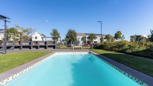 A swimming pool with a tiled edge, surrounded by a grassy area and residential buildings. - Neo-African apartment at the entrance of Durbanville wine route (Cape Town)