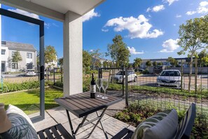 En udendørs patio med et bord, stole og en vinflaske, med udsigt over en parkeringsplads og bygninger.