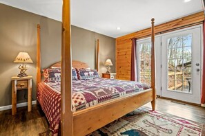 Uma cama com dossel adornada com um cobreleiro estampado, duas mesas de cabeceira com abajures, uma janela grande com vista para árvores e piso de madeira.