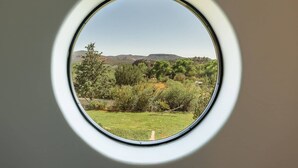 Ein rundes Fenster mit Blick auf eine grüne Landschaft, Bäume und ferne Hügel.