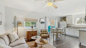Un salon moderne comprenant un canapé, une table basse et un fauteuil, un coin repas avec une table et des chaises, une cuisine avec des meubles blancs et des appareils électroménagers, et un ventilateur de plafond.