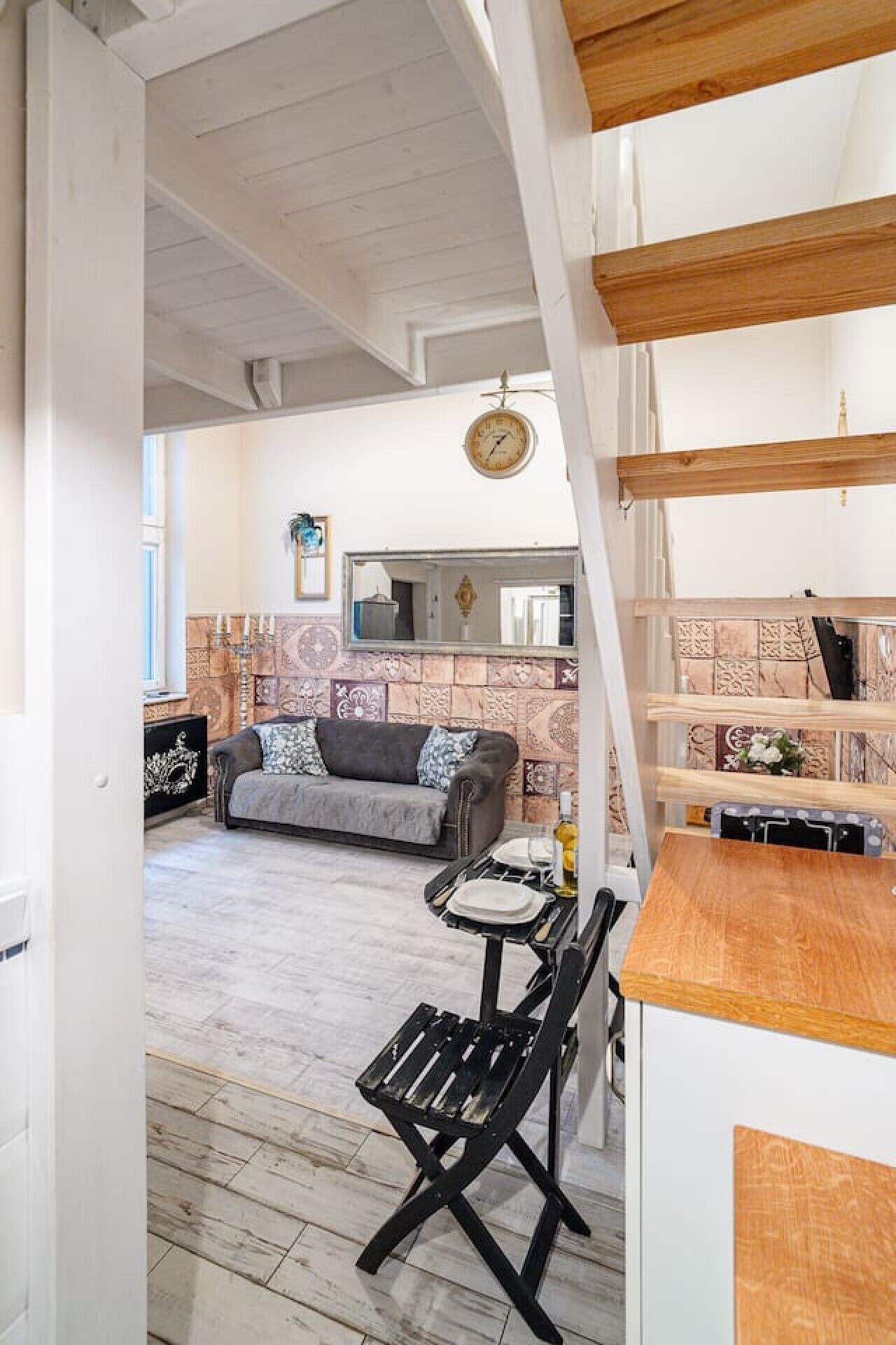 Een kamer met houten plafond, een bank, een eettafel met stoelen en een keukenhoek met een klok.