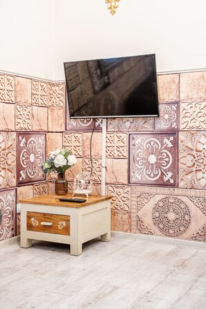 A room with a flat-screen TV mounted on the wall, a wooden side table with a vase of flowers, and decorative wall tiles. - Kraków Podgórze Incognito Studio — Wawel & Old Town Nearby (Kraków)