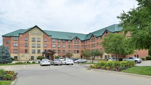 A Staybridge Suites hotel with a brick facade, green roof, and a parking lot with several cars.