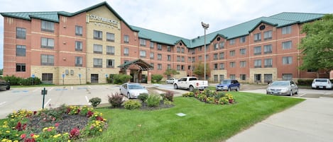 Un hotel Staybridge Suites con fachada de ladrillo, varios espacios de estacionamiento y un jardín ajardinado con flores coloridas.