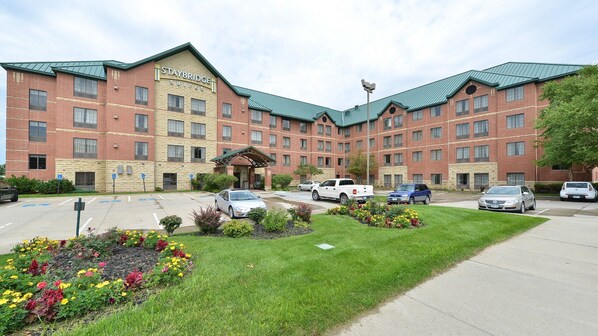 A Staybridge Suites hotel with a brick facade, multiple parking spaces, and a landscaped garden with colorful flowers.