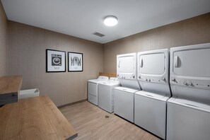 A laundry room with four white washing machines, two framed signs on the wall, and a wooden bench.