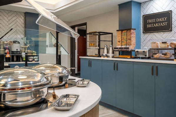 A breakfast buffet area with a sign indicating free daily breakfast, stainless steel food containers, a coffee machine, and a toaster.