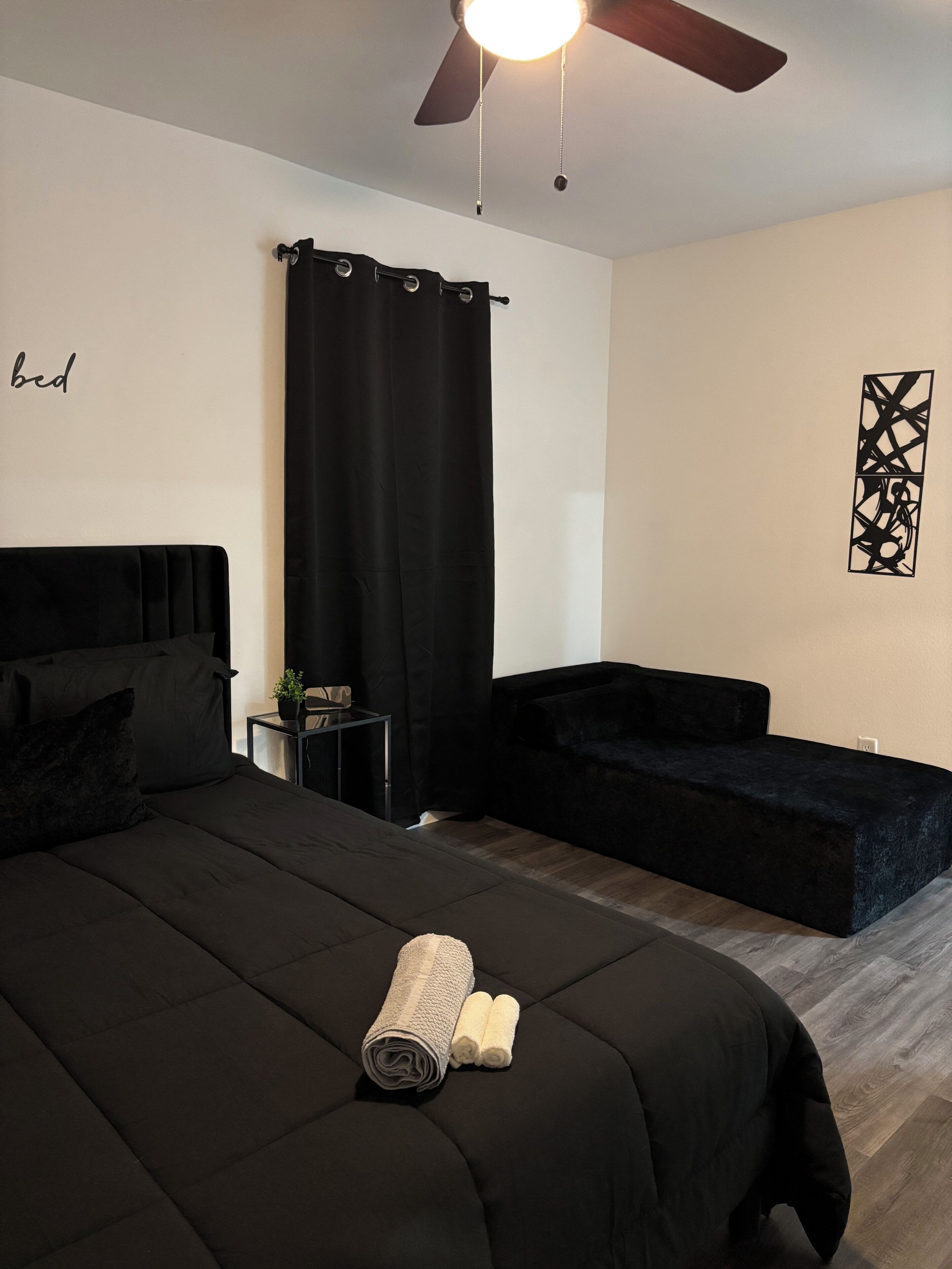 Un dormitorio con una cama, una mesita con una planta, un sofá negro, un ventilador de techo y una obra de arte colgada en la pared.