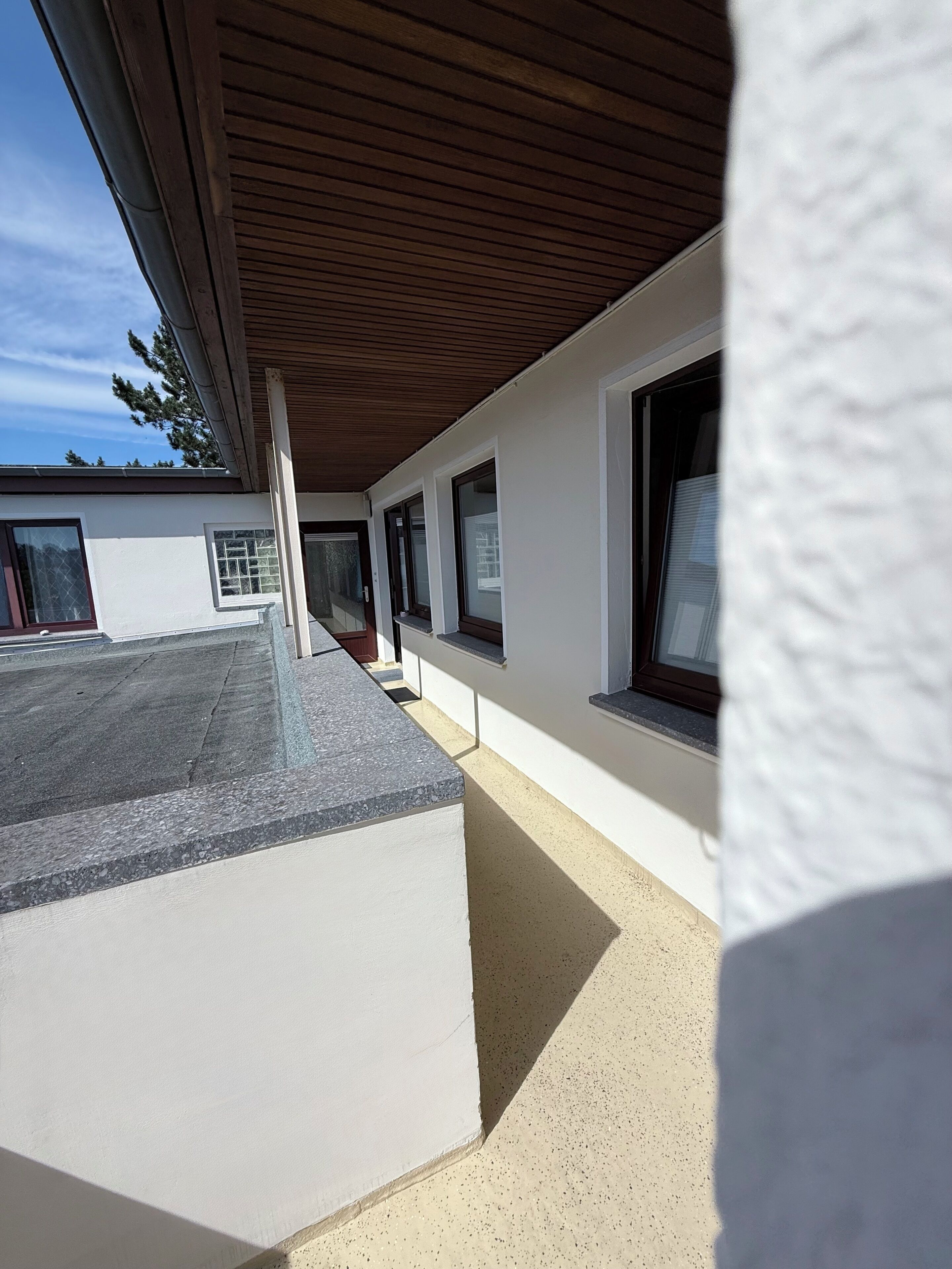 A covered outdoor area with a concrete floor, a wooden ceiling, and a white wall with windows.