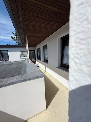 A covered outdoor area with a concrete floor, a wooden ceiling, and a white wall with windows.