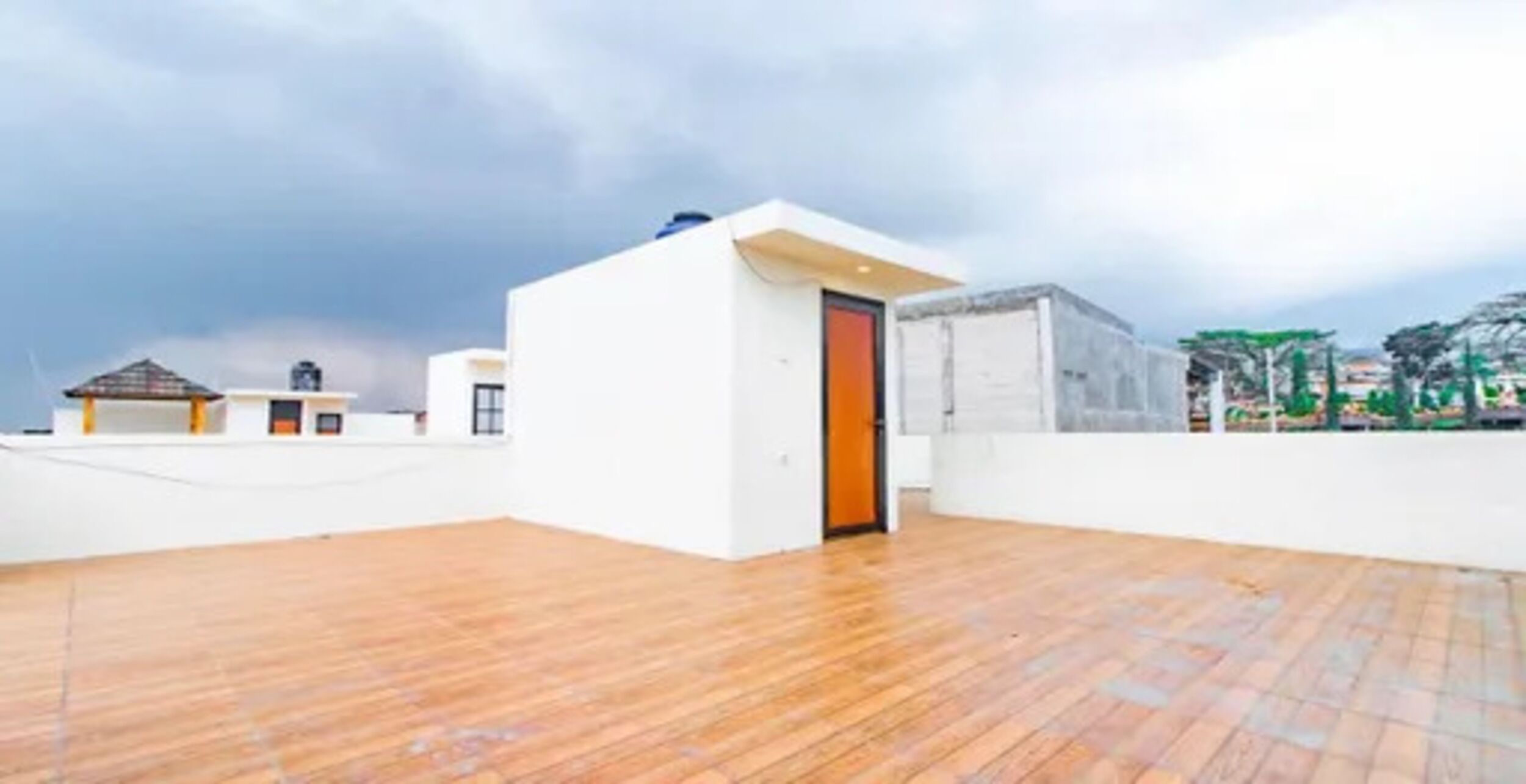 A spacious rooftop area with a wooden floor and a white building with an orange door.