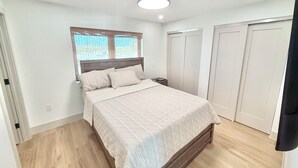 Un dormitorio con cama, mesita de noche, ventana con persianas y ventilador de techo.