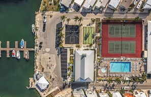 Luchtbeeld van een landgoed aan de kust met een steiger, tennisbanen, een zwembad en een gebouw met een schuin dak.
