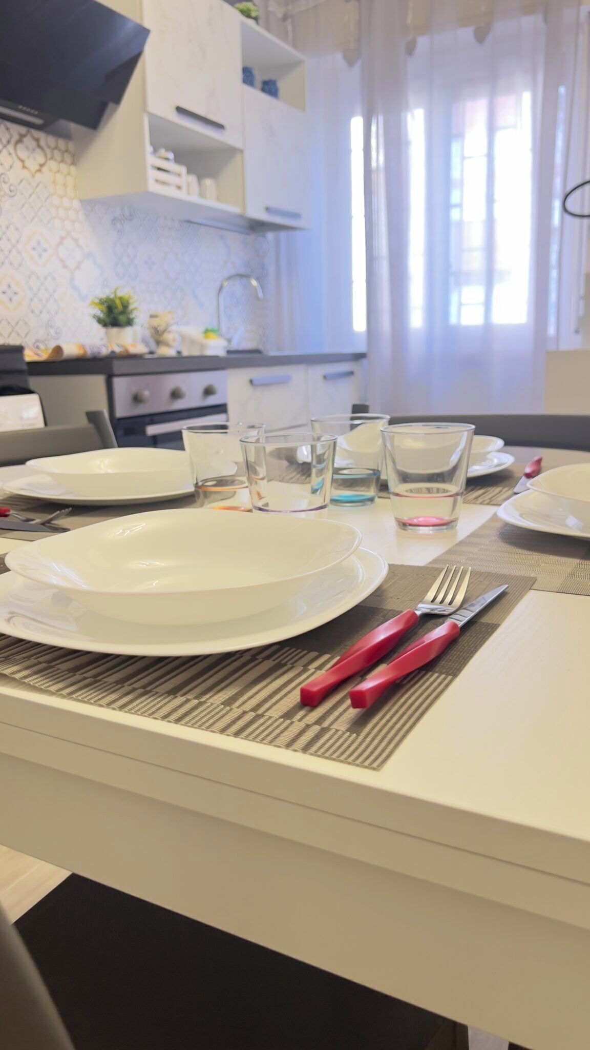 Une table à manger dressée avec des assiettes, des verres et des couverts, dans une cuisine moderne aux armoires blanches et au dosseret décoré.