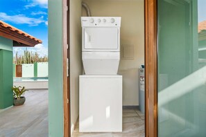 A compact, white Whirlpool washer and dryer unit in a small, enclosed laundry room with a view of a pool and cacti outside. - Alto Vista 107-h (Noord)