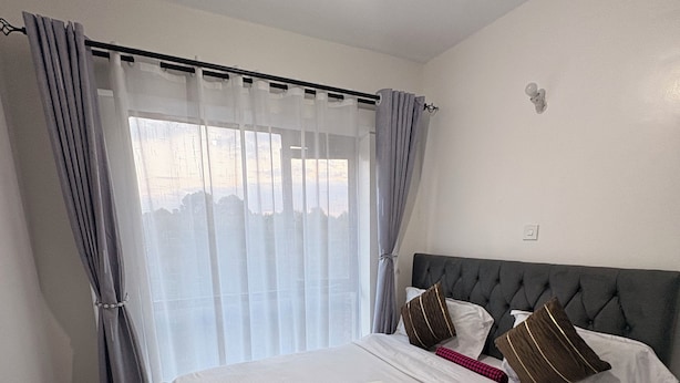 A hotel room with a large bed, curtains, a bedside table, and a nightstand. - Cozy one bedroom apartment (Ongata Rongai)
