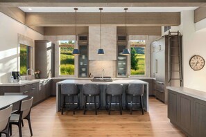 A modern kitchen with a central island, bar stools, a built-in oven, and a view of a green landscape through large windows. - Abode at White Clouds, hot tub, Sun Valley (Sun Valley)