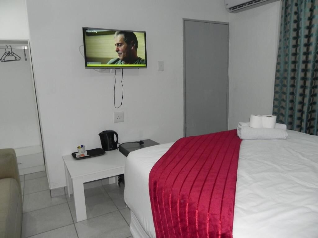 A hotel room with a bed, a TV mounted on the wall, a bedside table with a coffee maker, and a window with curtains.