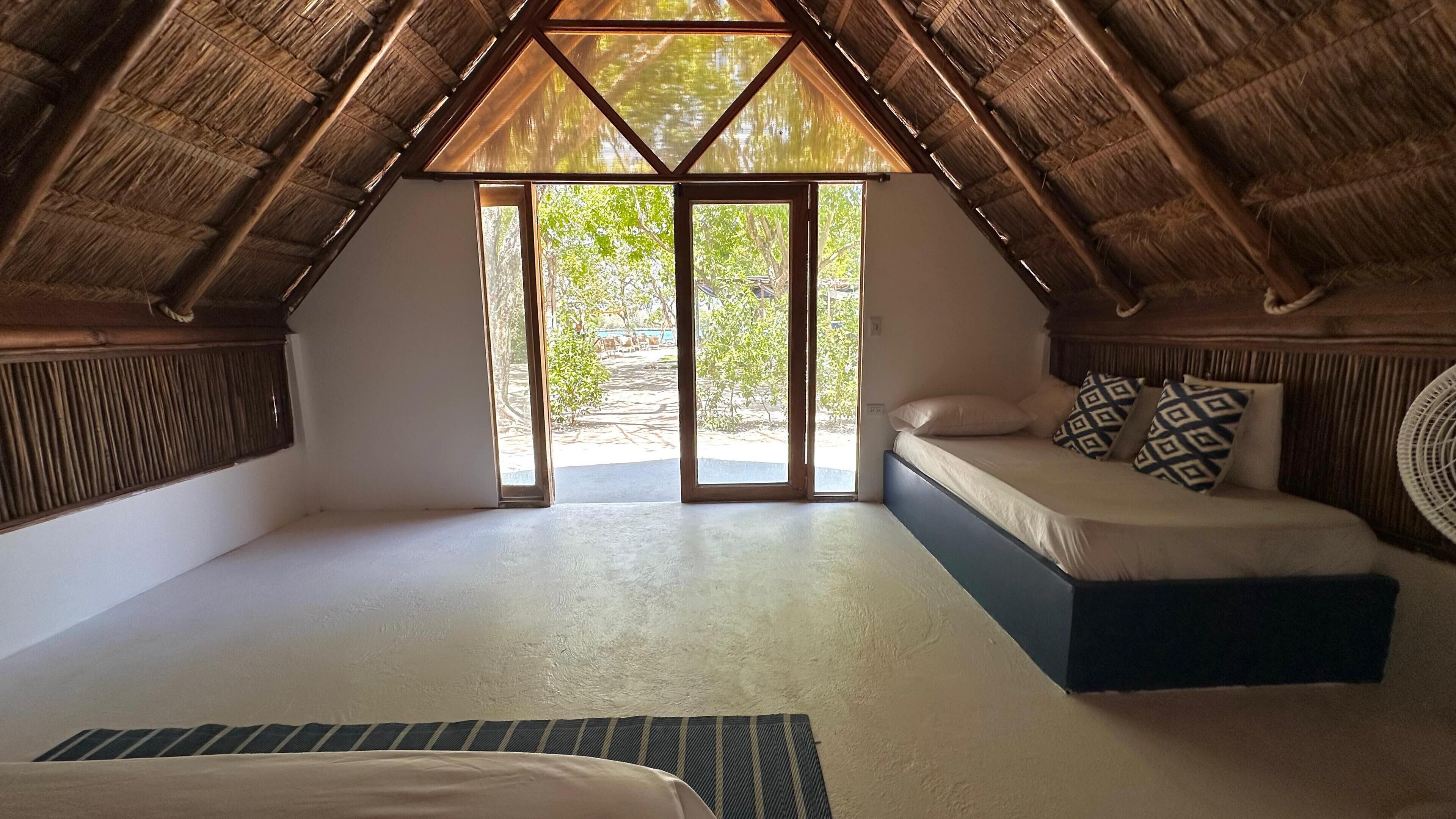 Una habitación con techo inclinado, una cama con almohadas estampadas y vistas al exterior a través de un ventanal.