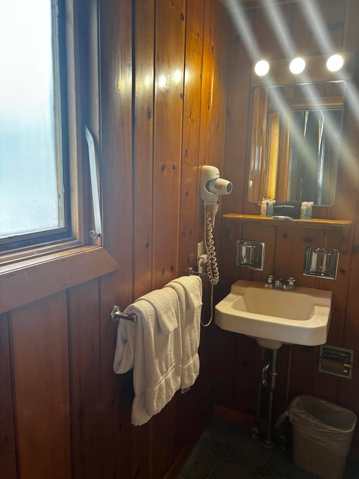 Een badkamer met houten wanden, een witte wastafel, een haardroger, een spiegel en een raam.