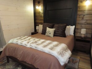 A bedroom with a bed, a wooden headboard, a nightstand with a lamp, and a wall-mounted light fixture. - Home Sweet Smoky (Pigeon Forge)