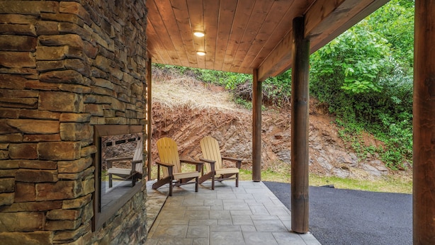 A covered outdoor seating area with stone walls and wooden beams, featuring Adirondack chairs and a stone fireplace. - Home Sweet Smoky (Pigeon Forge)