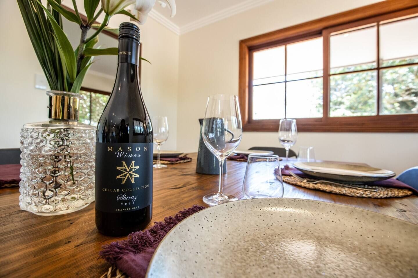 Ein Esstisch mit einer Flasche Mason Wines Shiraz 2016, einer Glasvase mit einer Pflanze und einem Fenster mit Blick auf Bäume.