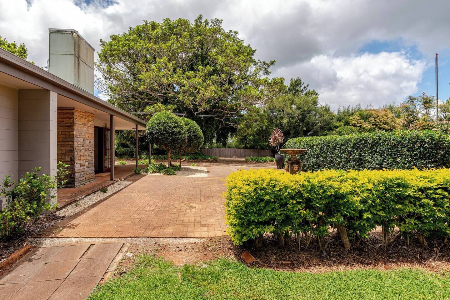 A residential property with a paved driveway, a covered outdoor area, and a well-maintained garden with various shrubs and a fountain.