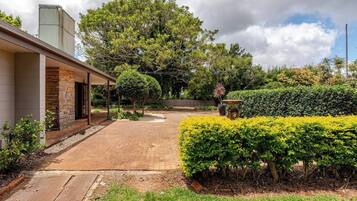 A residential property with a paved driveway, a covered outdoor area, and a well-maintained garden with various shrubs and a fountain.