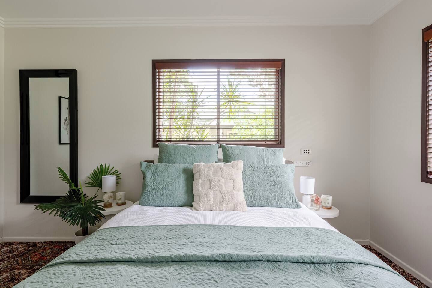 A bedroom with a bed, pillows, a bedside table, a mirror, and a window with blinds.
