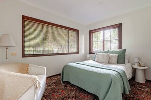 A bedroom with a bed, two bedside tables, a chair, and large windows with blinds. - Administration House Winery Stay (Tamborine Mountain)