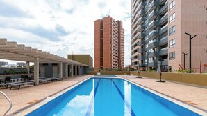 En swimmingpool med et pergolaoverdækket område og liggestole, omgivet af høje bygninger.