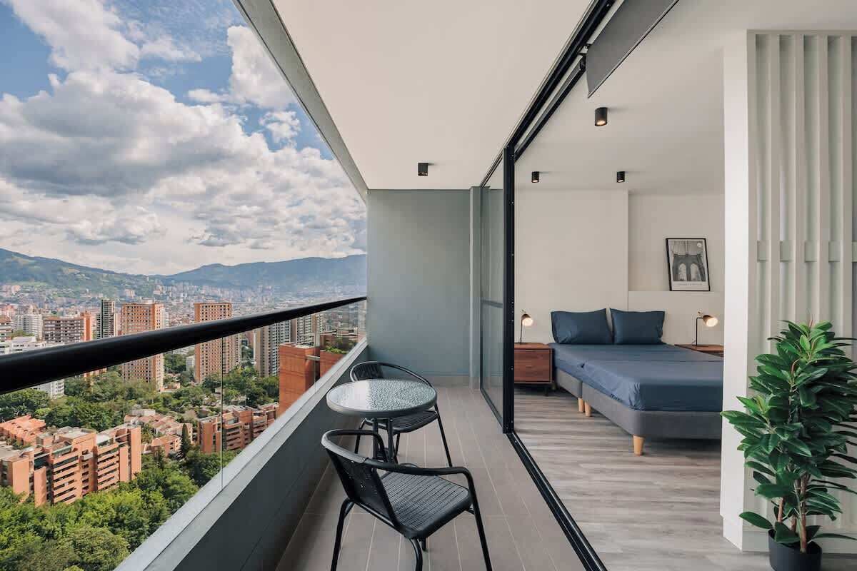 A balcony with a table and chairs, a bed with a blue mattress, and a city view.