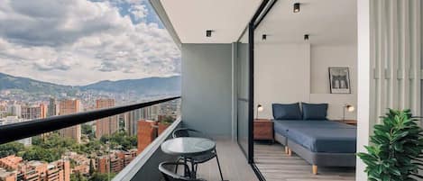 A balcony with a table and chairs, a bed with a blue mattress, and a city view.