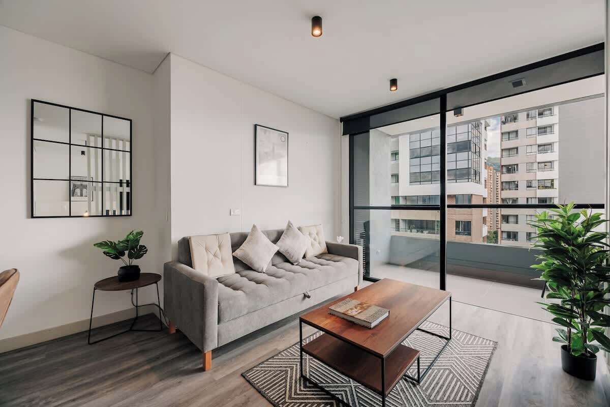 A modern living room with a grey sofa, a wooden coffee table, and a large window offering a view of buildings.