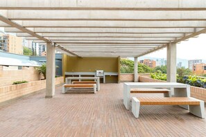 Een terras buiten met betonnen banken en tafels, een barbecue en uitzicht op omliggende gebouwen.