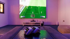 Ein Zimmer mit großem Bildschirm, der ein Sportspiel zeigt, einer Spielkonsole und einer gemütlichen Sitzecke.