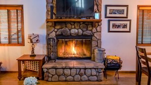 A stone fireplace with a television above it, flanked by windows with blinds. - Cozy Cabin Near Snow Summit w Loft & Fireplace (Big Bear Lake)