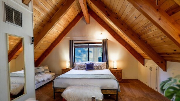 A bedroom with a sloped ceiling, a bed with white bedding, a window with curtains, and a small bedside table with a lamp. - Cozy Cabin Near Snow Summit w Loft & Fireplace (Big Bear Lake)