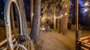 A rustic outdoor seating area with wooden benches and chairs, surrounded by trees and string lights. - Cozy Cabin Near Snow Summit w Loft & Fireplace (Big Bear Lake)