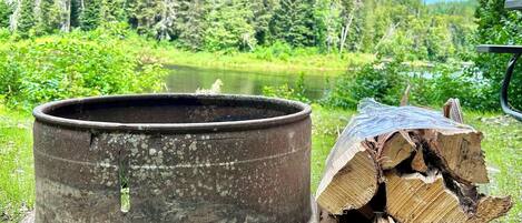 A metal container and a stack of firewood by a lake with trees in the background.