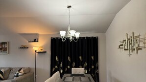 Spisestue med bord, stole og en lysekrone. En sofa og en gulvlampe ses i baggrunden.