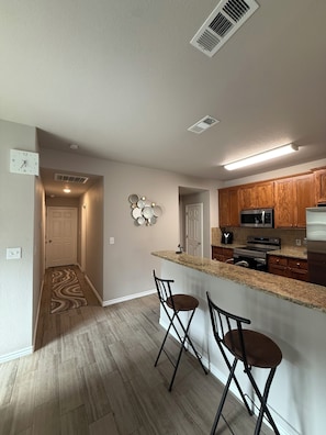 A kitchen with wooden cabinets, a microwave, and a refrigerator. There are two bar stools and a clock on the wall.