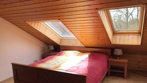 Une chambre avec un plafond en pente, un lit recouvert d’une couvre-lit rose et blanc, deux tables de chevet avec des lampes et une lucarne.