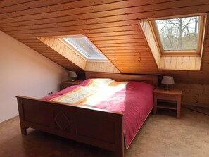 Ein Schlafzimmer mit schrägverdeckter Decke, einem Bett mit rosa-weißer Bettdecke, zwei Nachttischen mit Lampen sowie einem Oberlicht.