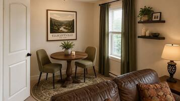 A living room with a brown leather couch, a round wooden table with two chairs, a framed picture on the wall, a window with curtains, and a shelf with decorative items.