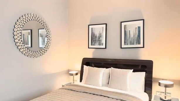 Un lit bien fait, recouvert d’une courtepointe matelassée, un miroir rond avec un cadre décoratif, deux tableaux encadrés accrochés au mur et des lampes de chevet.