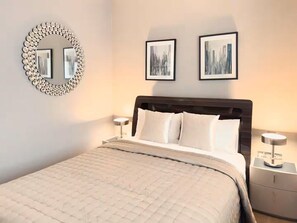 Ein ordentlich bezogenes Bett mit einer gesteppten Decke, ein runder Spiegel mit dekorativem Rahmen, zwei gerahmte Bilder an der Wand und Nachttischlampen.